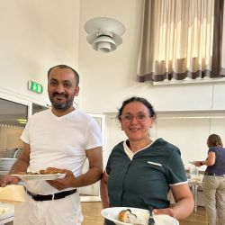 Zwei Personen stehen lächelnd in einem hellen Raum und halten Teller mit Essen in den Händen.