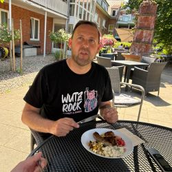 Ein Mann im "Wutz Rock"-Shirt genießt im Freien an einem Tisch ein Gericht mit Fleisch und Salat.
