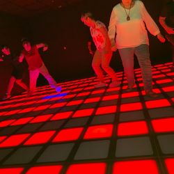 Mehrere Personen tanzen auf einem rot leuchtenden Boden in einem dunklen Raum, während farbige Lichter die Stimmung heben.