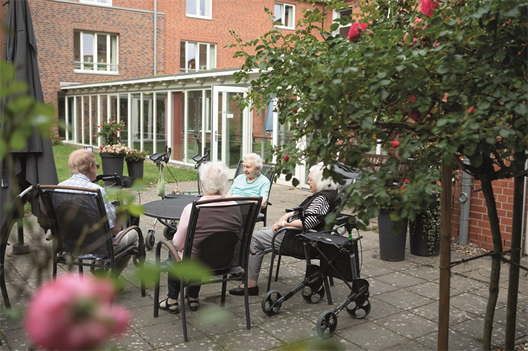 Ältere Menschen sitzen gemütlich im Garten eines Seniorenheims und unterhalten sich entspannt an einem Tisch.