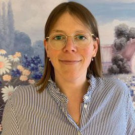 Eine Frau mit Brille lächelt vor einem idyllischen Hintergrund mit Blumen und einem Haus, was an einen Garten erinnert.