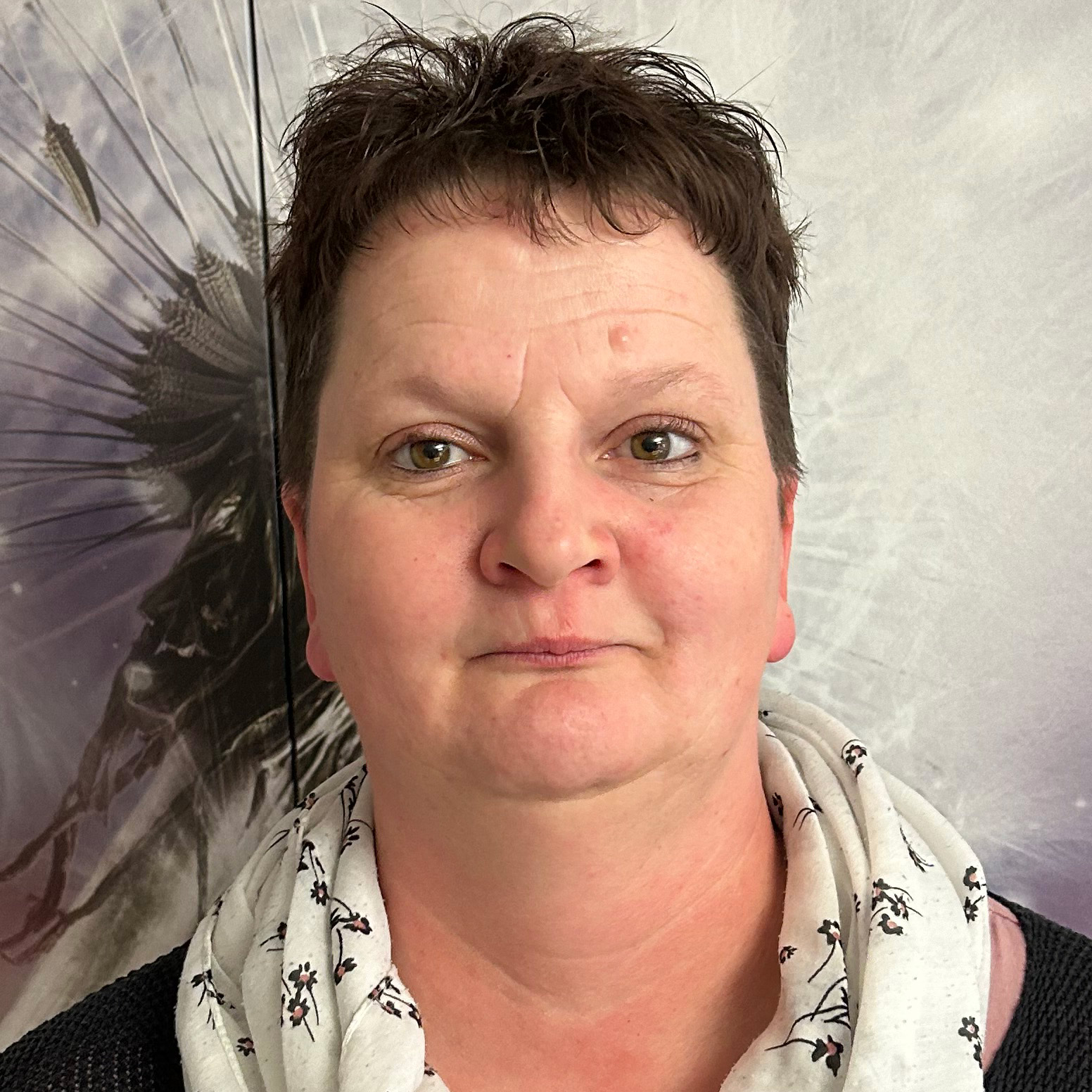 Eine Frau mit kurzem Haar und gemustertem Schal steht vor einem kunstvoll gestalteten, detailreichen Hintergrund.