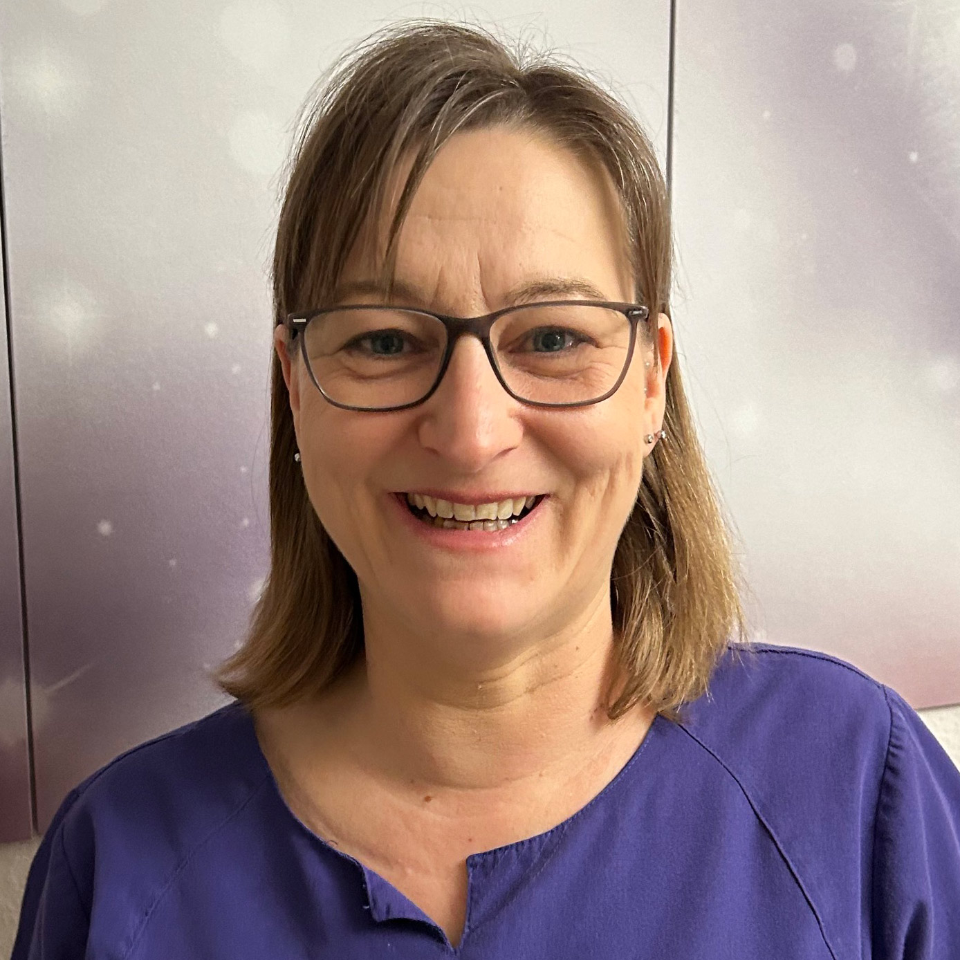 Eine lächelnde Frau mit Brille trägt ein lila Oberteil vor einem unscharfen, hellen Hintergrund.