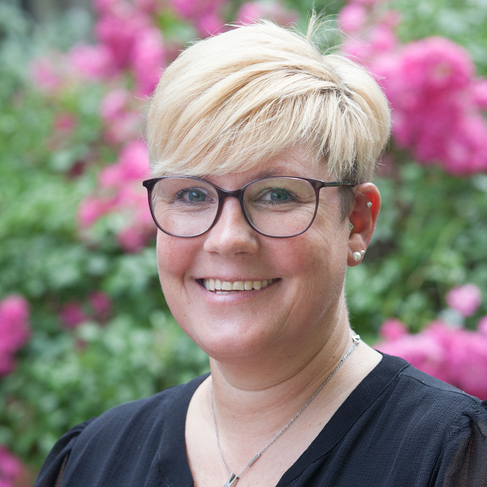 Eine lächelnde Frau mit Brille und kurzem blondem Haar steht vor einem Hintergrund aus blühenden, rosa Blumen.