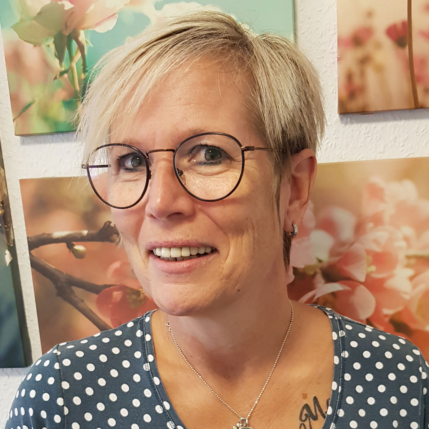 Eine Frau mit Brille und kurzen Haaren lächelt vor einem blumigen Hintergrund, der an ein Werk von Michaela Bittner erinnert.