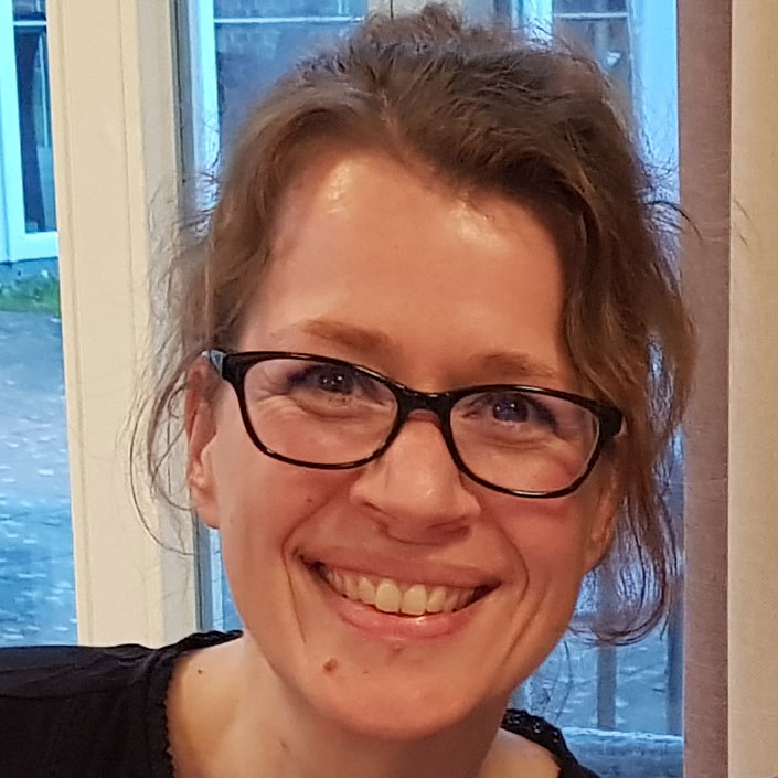 Eine lächelnde Frau mit Brille steht vor einem Fenster, durch das ein grauer Tag sichtbar ist.