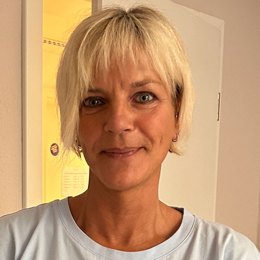 Eine lächelnde Frau mit kurzem, blondem Haar steht in einem hellen Raum und strahlt Ruhe und Freundlichkeit aus.