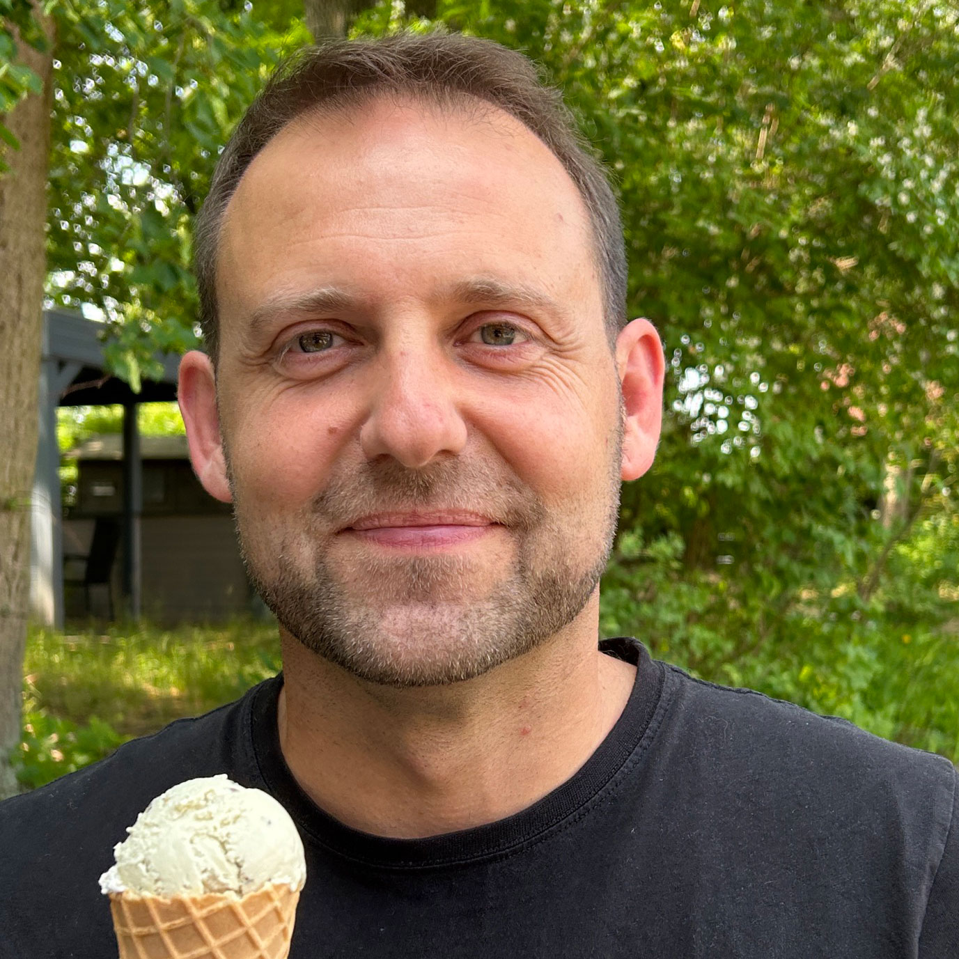 Ein Mann lächelt glücklich, während er ein Eis in der Hand hält, umgeben von einer grünen, natürlichen Umgebung.