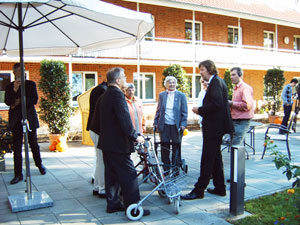 Eine Gruppe von Menschen unterhält sich entspannt bei sonnigem Wetter auf einer Terrasse vor einem Backsteingebäude.