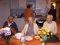 Drei ältere Menschen genießen die Betreuung in einer gemütlichen Küche mit Blumen auf dem Tisch.