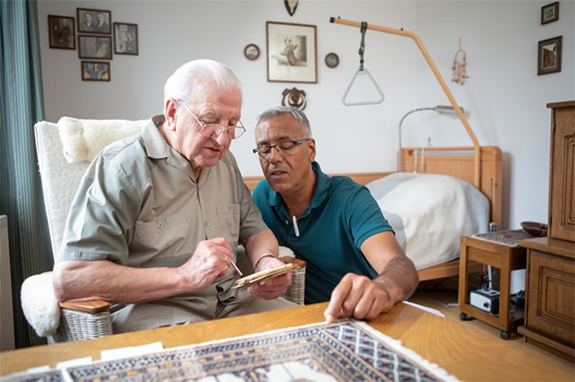 Ein älterer Mann und sein Betreuer betrachten ein Notizbuch im gemütlichen Wohnzimmer neben einem Bett.