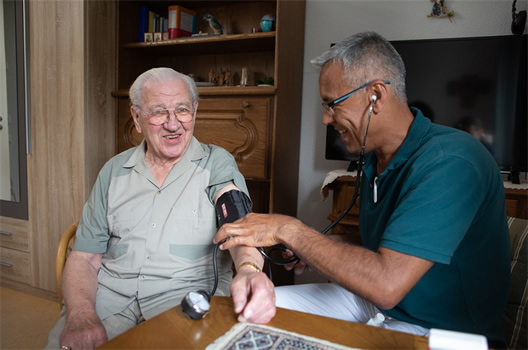Ein älterer Mann lächelt, während ein Arzt in einem gemütlichen Raum seinen Blutdruck mit einem Messgerät überprüft.