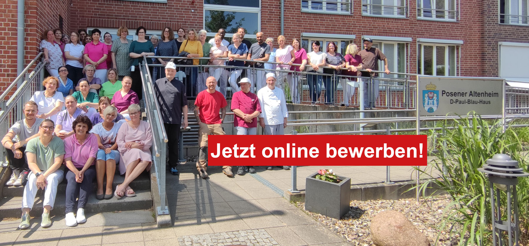 Eine große Gruppe von Mitarbeitern posiert fröhlich vor einem Altenheim mit einem Schild "Jetzt online bewerben!".
